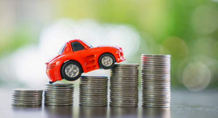 A small red toy car sits on top of coins ascending from left to right, with blurred greenery in the background.