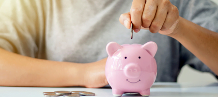 A person places a coin into a piggy bank using their left hand. Spare coins sit to the right of the piggy bank.