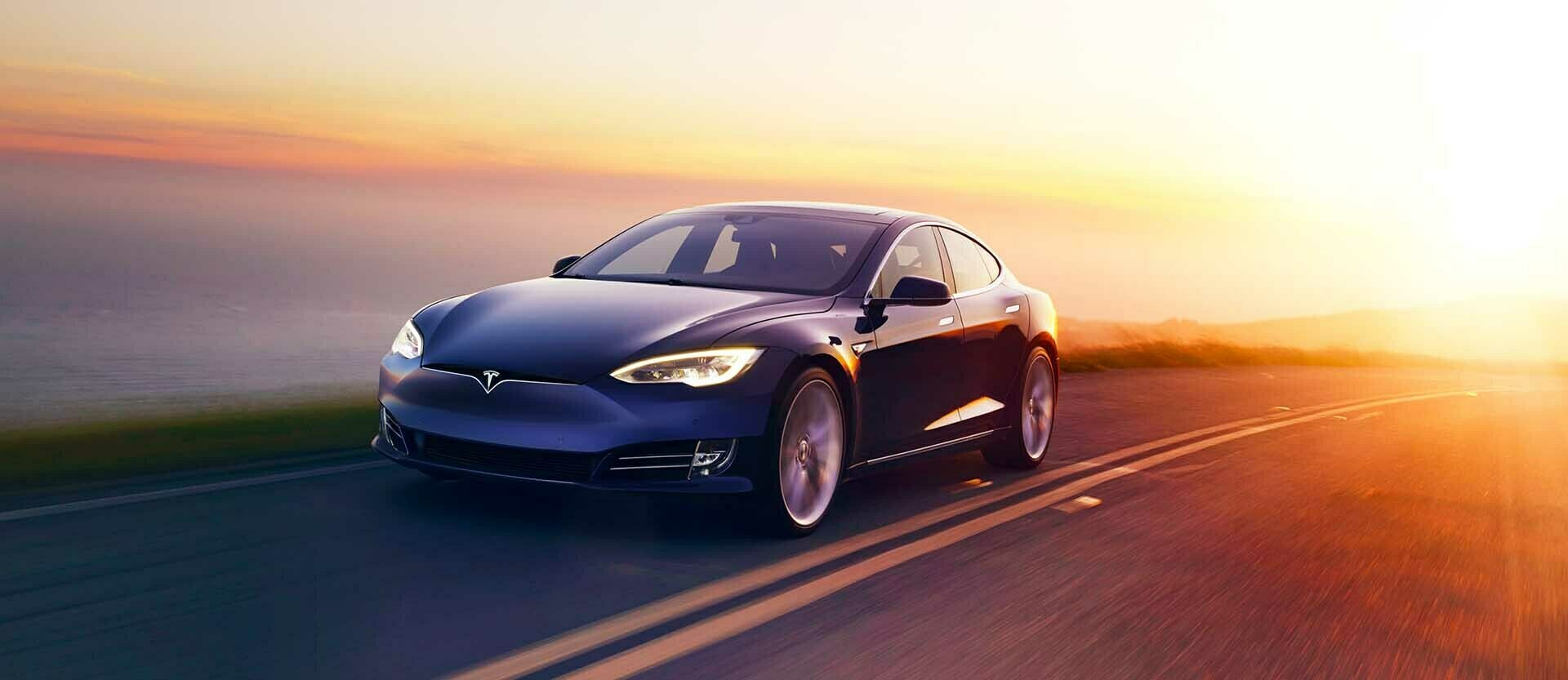 A blue Tesla Model S on an open road with the sea to its right and bright sunlight behind it to the left.