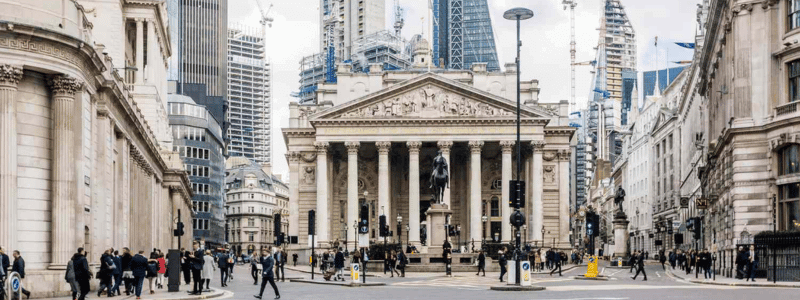 The front of the Bank of England, taken in day time with pedestrians in shot and London skyscrapers in the background.