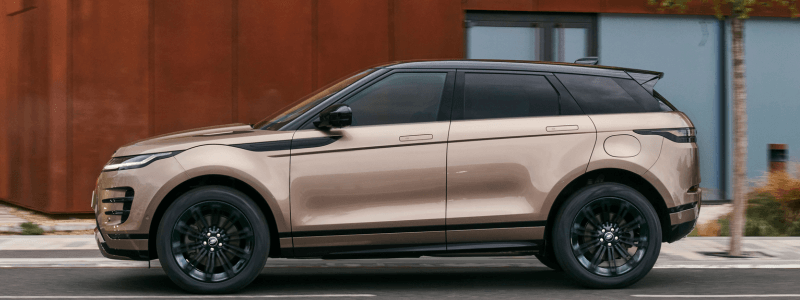 A side view of a bronze Range Rover Evoque driving on a road, with a building in the backdrop.