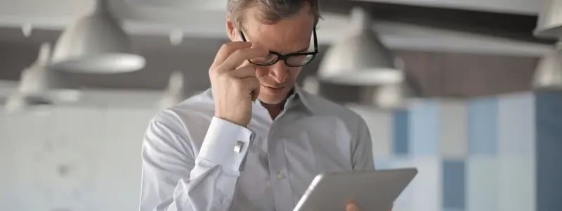 A business person adjusting their glasses whilst looking at a tablet device, situated in an office setting.