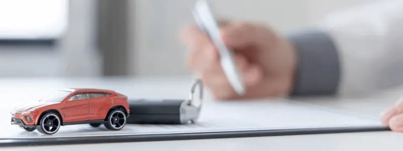 A small model car and car keys sit on a document, whilst a person signs, representing a car purchase.