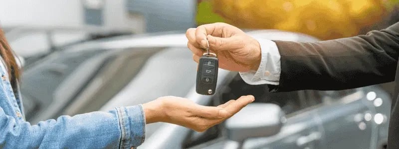 Car keys being handed to someone, with a car in the background, likely showing a car purchase.