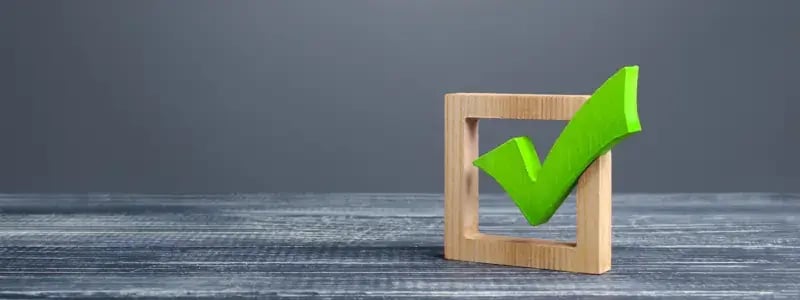 A green tick emerging out of a hollow wooden square, against a dark grey background.