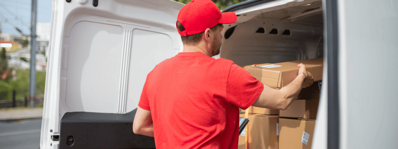 A man in a red hat and polo shirt loads boxes into a van, representing a delivery driver.