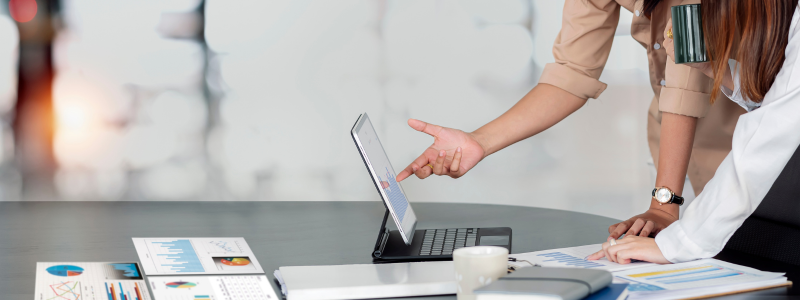 A person points to a tablet device with multiple analysis documents spread around a table, whilst someone else engages.