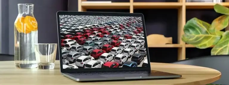 A desk set up with cupboards in the background. On the desk is a large jug of water with orange slices in, next to an empty glass and an open laptop displaying rows of cars, symbolising an online car market.