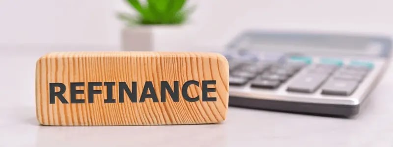 A wooden block with 'REFINANCE' written on it, with a calculator and plant in the background, signalling a place of business/work.