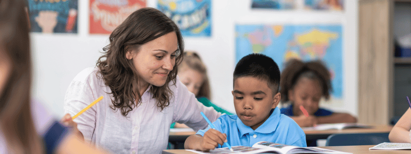 A female teacher helping a young student in a classroom setting.