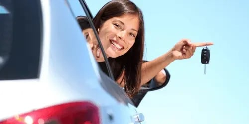 A woman leaning out of a car window and looking back, whilst holding car keys up and smiling.
