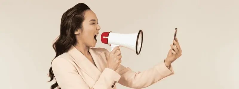 A side on shot of a business woman shouting into a megaphone whilst holding a mobile device slightly ahead of the megaphone.