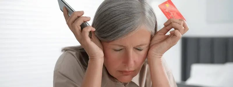 A woman with her hands by the sides of her head in visible distress, whilst holding a credit card and a phone, symbolising money struggles.