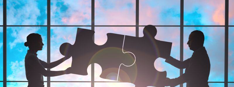Silhouettes of two people piecing together larger jigsaw pieces in an office setting, representing business partnerships.