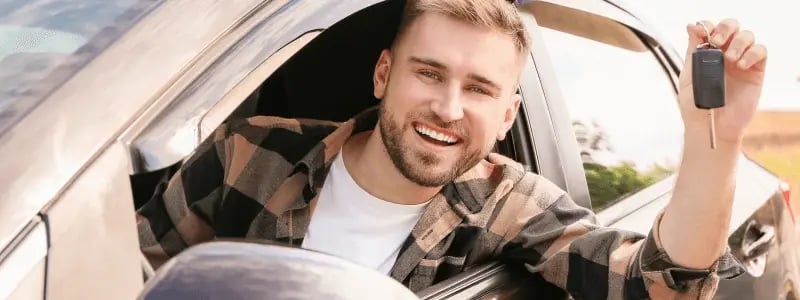 A person smiles whilst holding car keys from his car window, likely representing a new car purchase.