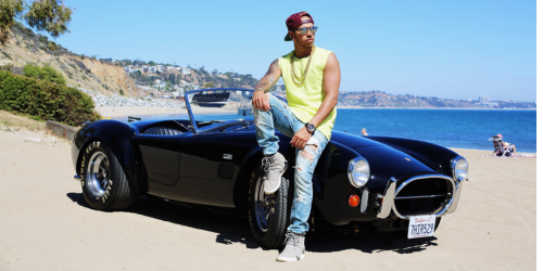 Lewis Hamilton posing in front of his Shelby Cobra, situated on a beach in summer time.