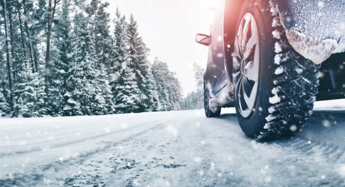 A car from the rear left tyre at ground level, with snow on the road and tyre and snow covered trees along the left hand side of the road.
