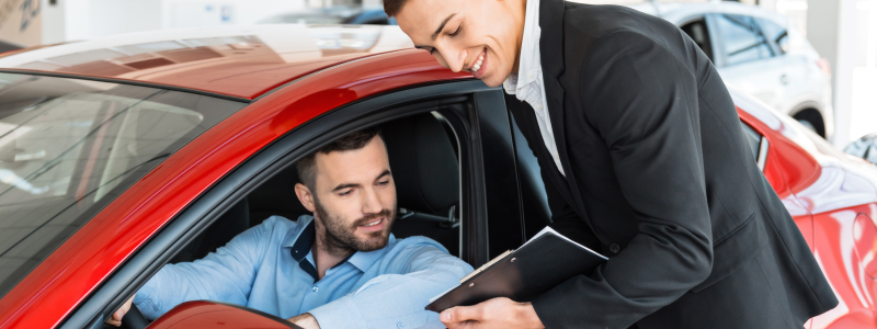 A person sat in a car is shown something on a tablet device by a car salesman stood next to the car.