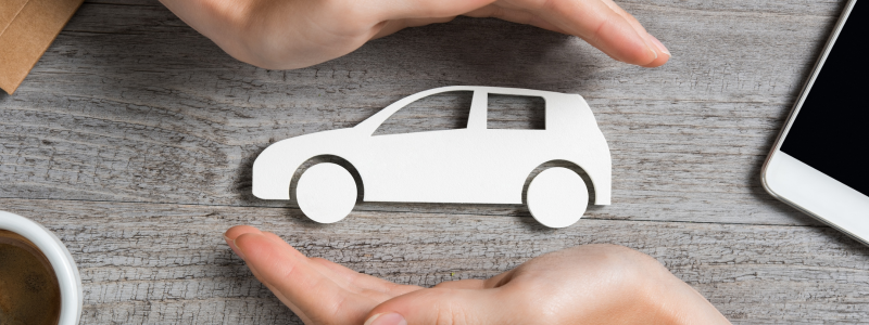 Hands holding a paper cutout of a car over a wooden desk, suggesting the concept of car insurance or vehicle protection.