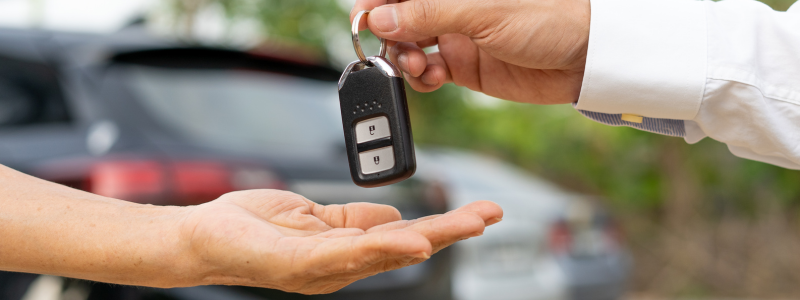 A person hands car keys to another person with a car in the background.
